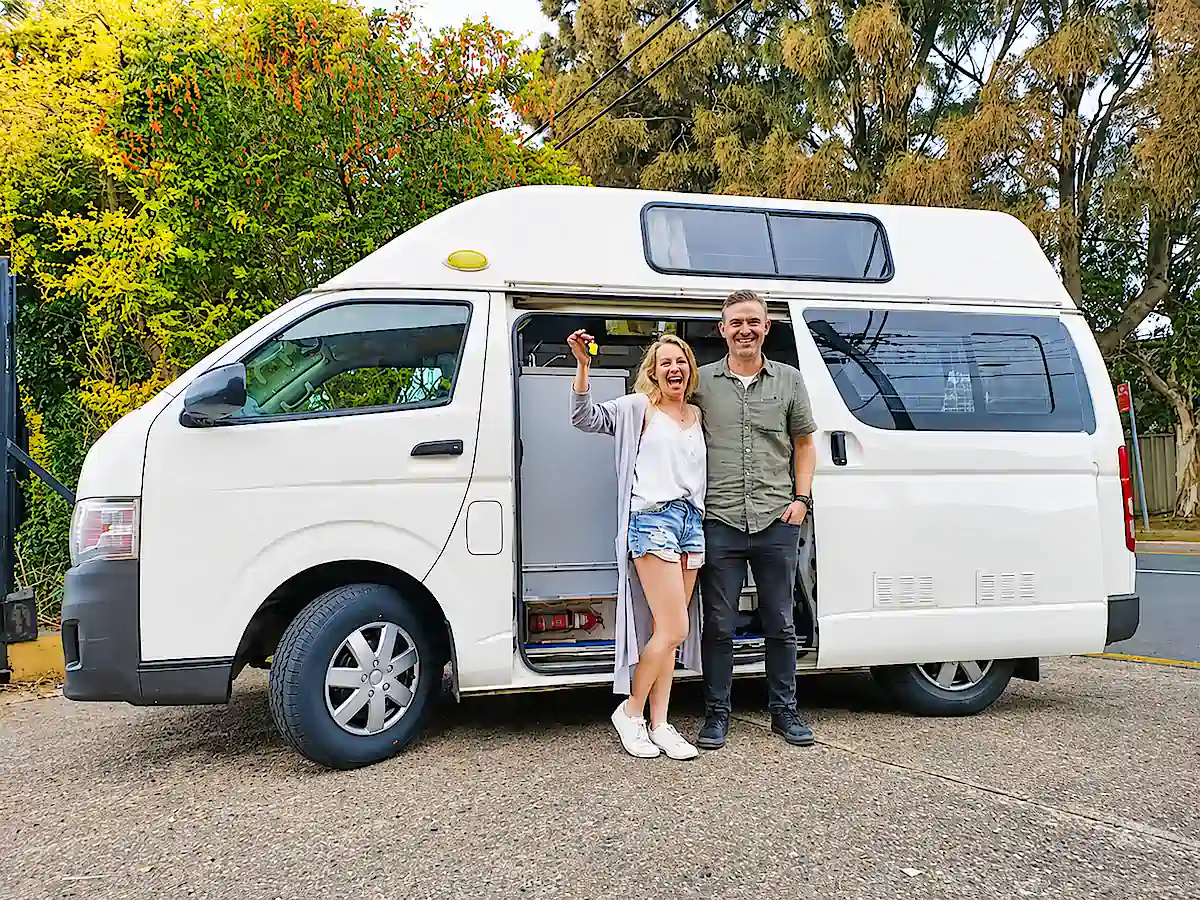 Couple de voyageurs heureux devant leur campervan Toyota après l’achat chez Travelwheels, avec les clés du véhicule en main avant leur road trip en Australie et l’option de rachat garanti pour la fin du voyage.